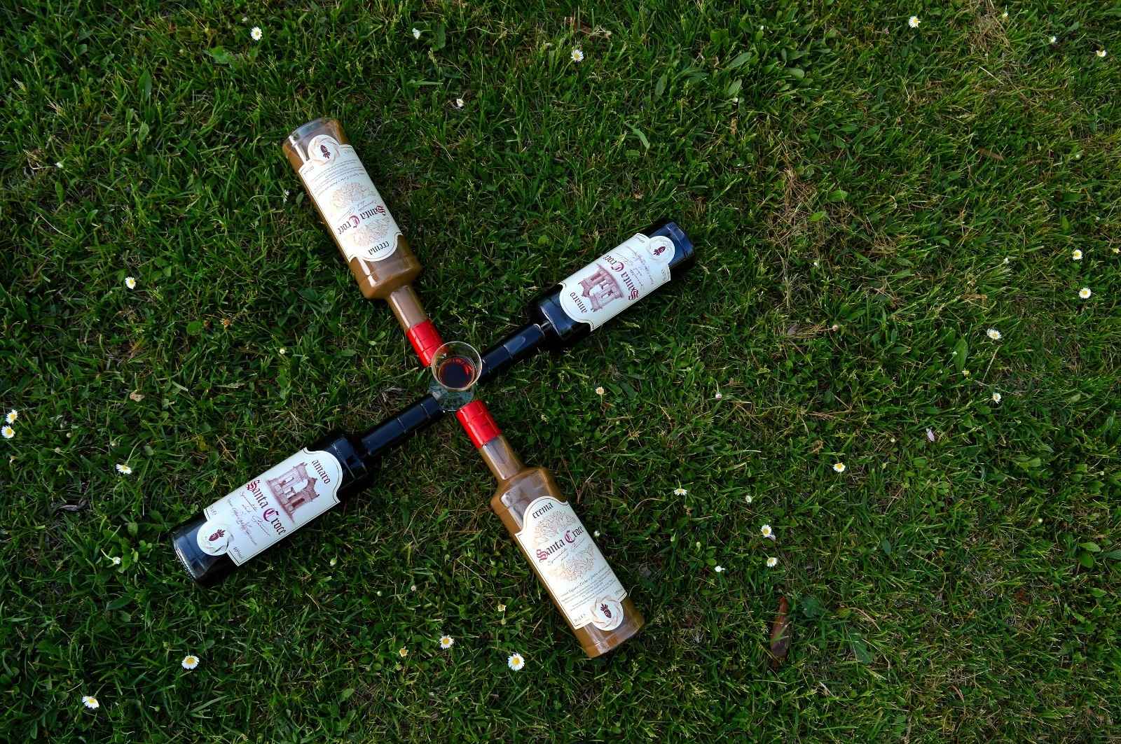 Bicchiere di Amaro Santa Croce al centro, circondato da quattro bottiglie disposte a raggiera su prato verde, evocando un’esperienza di gusto artigianale italiana